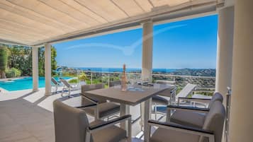 A covered outdoor seating area with a pool, chairs, and a table with wine glasses.