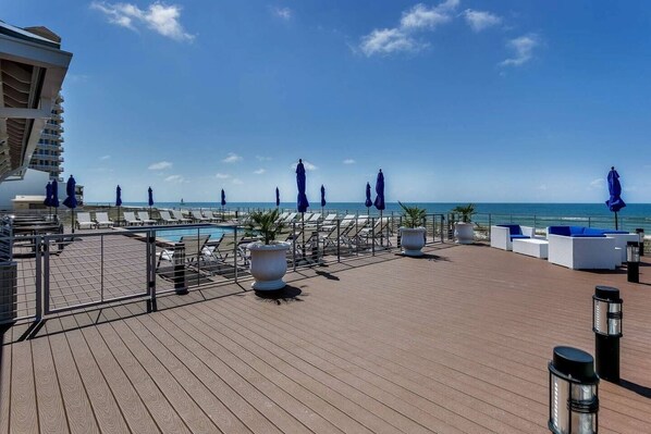 A rooftop pool area with lounge chairs and umbrellas, overlooking the ocean.