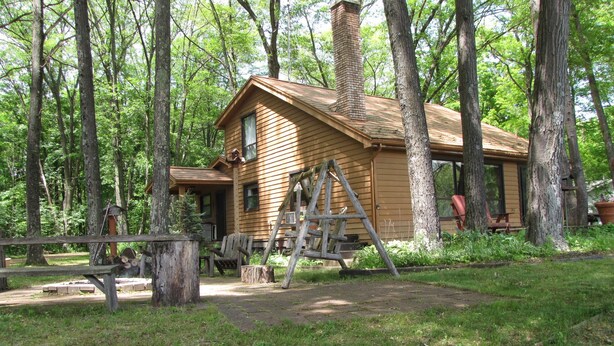 A rustic cabin with a chimney, surrounded by tall trees and a wooden swing set. - Nature lovers retreat right on the edge of the river (Hersey)