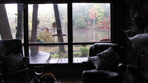 A room with a large window overlooking a lake, wooden deck, and outdoor furniture. - Nature lovers retreat right on the edge of the river (Hersey)