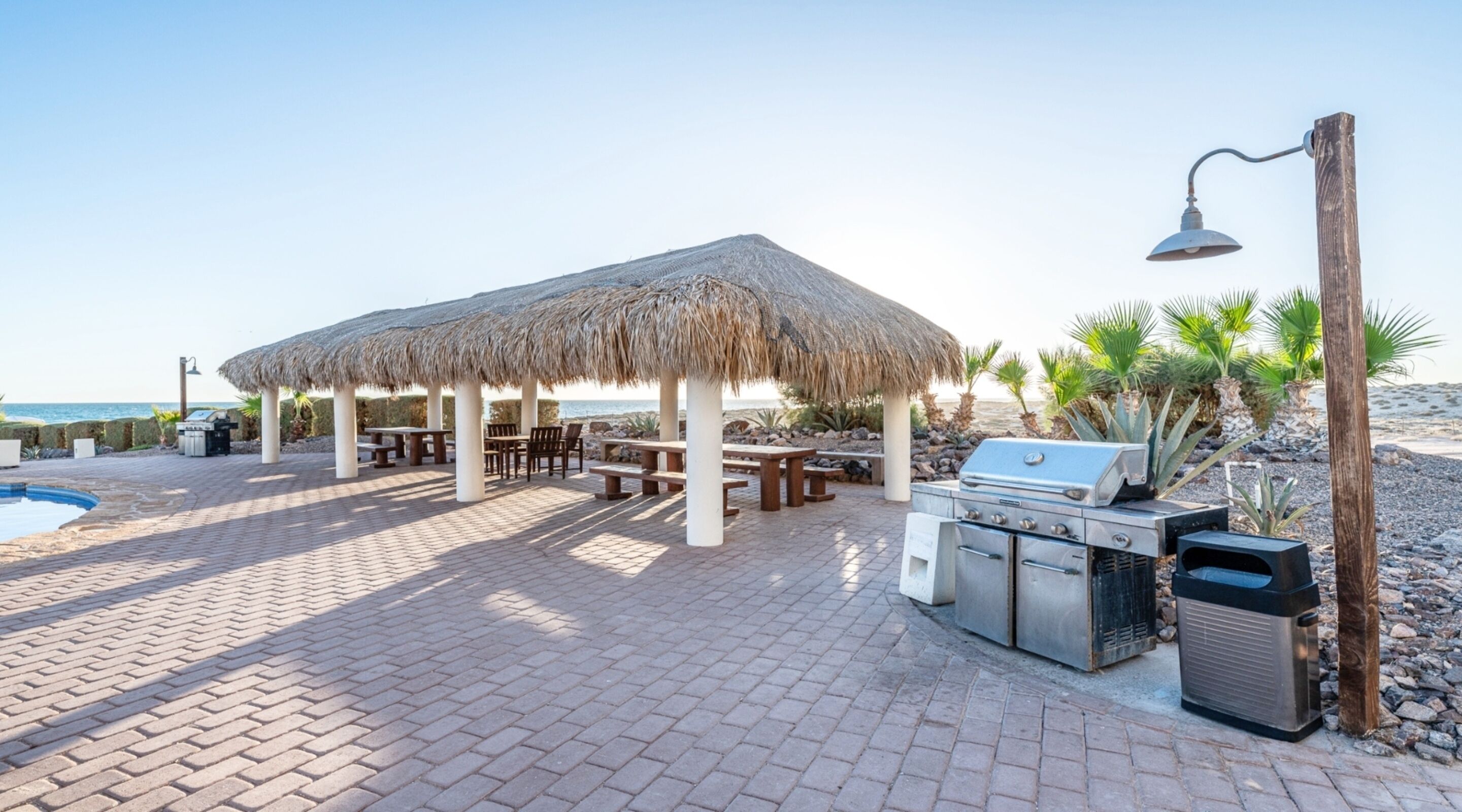 Een terras aan het strand met een gazebopje met rieten dak, een buitenbarbecue en een zitgedeelte.
