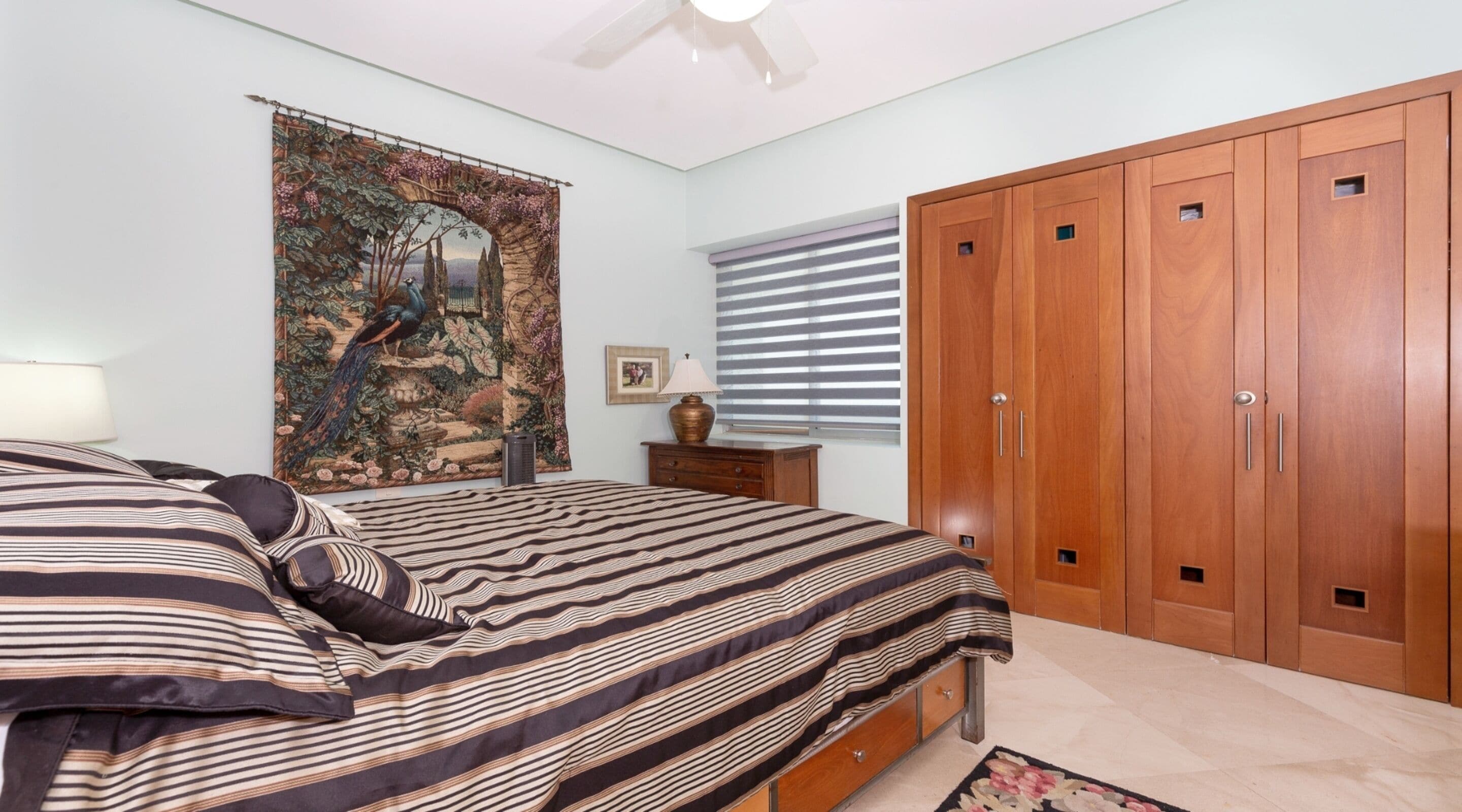 A bedroom with a bed, a nightstand, a lamp, a tapestry, a window with blinds, and a wardrobe.