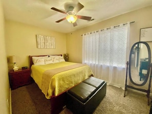 Une chambre à coucher comprenant un lit, un ventilateur de plafond, une table de chevet, un miroir et une fenêtre avec des stores.