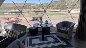 A room with a view of a hillside, two armchairs, a small table with a clock, and a red picnic table outside. - Grand Canyon Glamping Resort (Meadview)