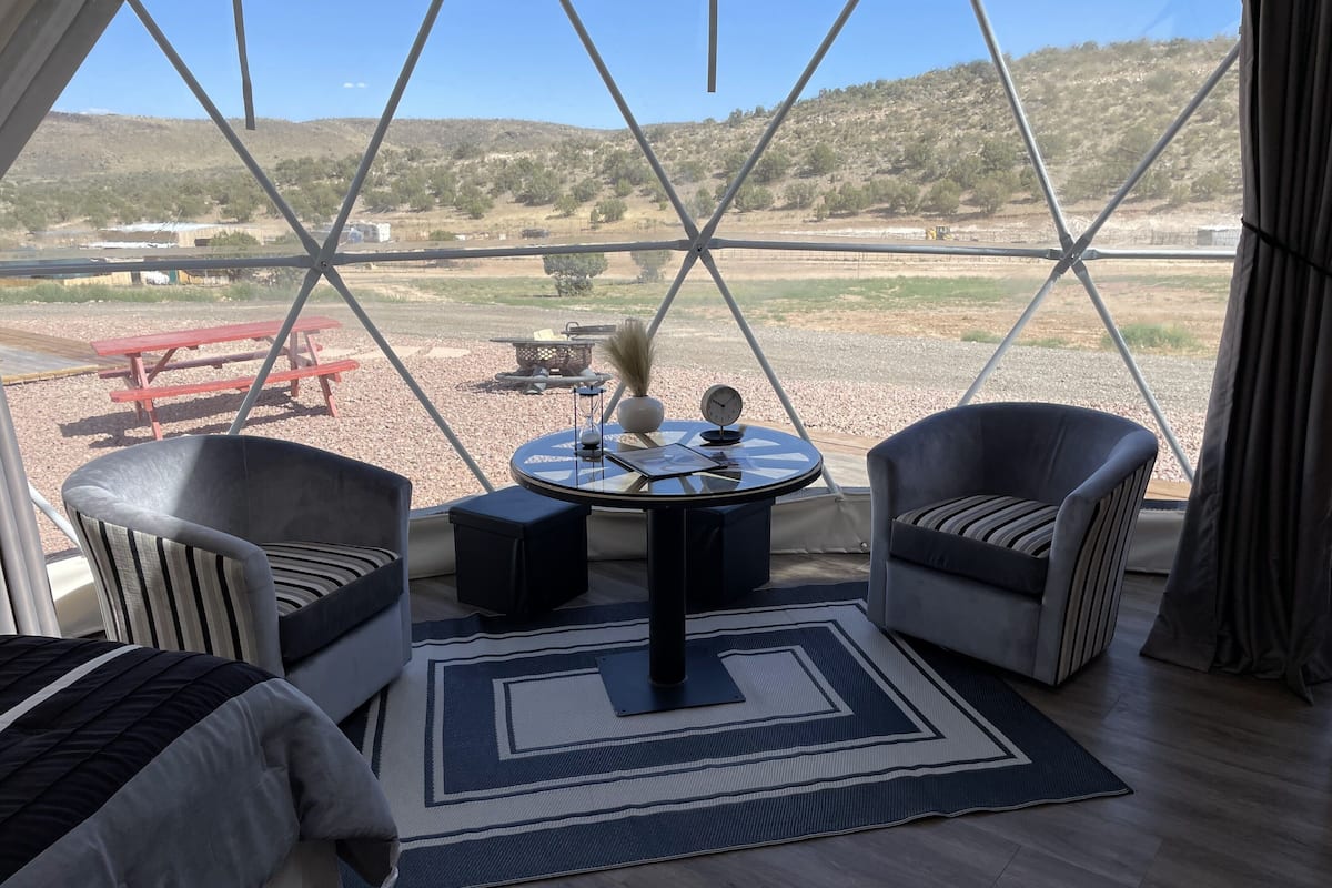 A room with a view of a hillside, two armchairs, a small table with a clock, and a red picnic table outside.