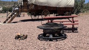A covered wagon with a wooden structure, a picnic table, and a fire pit in a gravel area. - Grand Canyon Glamping Resort (Meadview)