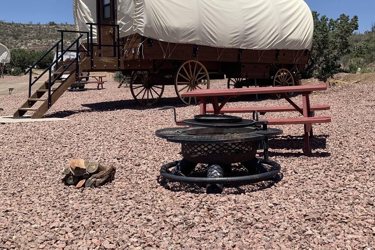 A covered wagon with a wooden structure, a picnic table, and a fire pit in a gravel area.