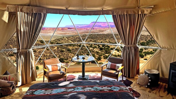 A tented room with a bed, two chairs, a small table, and a view of a desert landscape. - Grand Canyon Glamping Resort (Meadview)