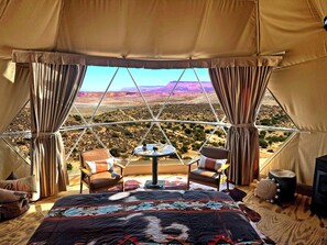 A tented room with a bed, two chairs, a small table, and a view of a desert landscape. - Grand Canyon Glamping Resort (Meadview)