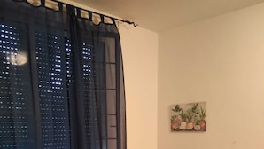 Un dormitorio con cama, una silla, un escritorio con lámpara, una ventana con cortinas y una planta en la pared.