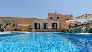 A swimming pool with clear blue water, surrounded by a building with a tiled roof and a stone wall. There are lounge chairs and umbrellas around the pool area.