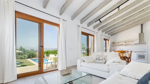 A modern living room with a glass coffee table, white sofas, and a kitchen area visible through an open sliding door.