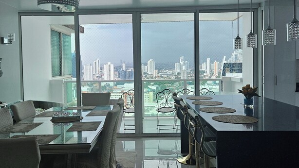 Ein modernes Wohnzimmer mit einem Glastisch, bequemen Sitzgelegenheiten und Blick auf eine Skyline durch große Fenster.