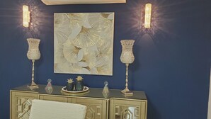 Ein Zimmer mit dunkelblauer Wand, einem dekorativen Gemälde, zwei Kristallvasen und einem Spiegelkorpus mit weißem Sessel.