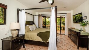 A four-poster bed with a canopy, a television mounted on the wall, and a balcony with a view of greenery. - Ocean-View Condo / Lagoon Pool & Swim-Up Bar (Tamarindo)