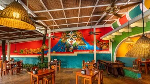 A restaurant interior with wooden tables and chairs, a mural on the wall, and hanging lights. - Ocean-View Condo / Lagoon Pool & Swim-Up Bar (Tamarindo)