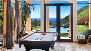 Ein Billardtisch in einem Raum mit großen Fenstern und Blick auf Pool und Landschaft.