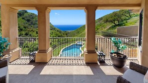 Ein Balkon mit Blick auf ein Schwimmbecken und das Meer, umgeben von Grünpflanzen und Säulen.