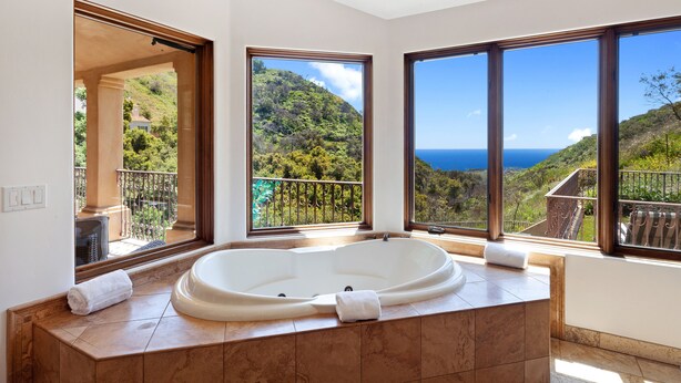 Ein Badezimmer mit einer großen Badewanne und zwei großen Fenstern mit Blick auf eine Berglandschaft und das Meer.