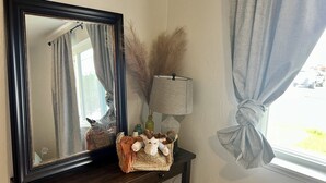 Un miroir avec un cadre foncé, une lampe, un panier décoratif et une fenêtre avec des rideaux.