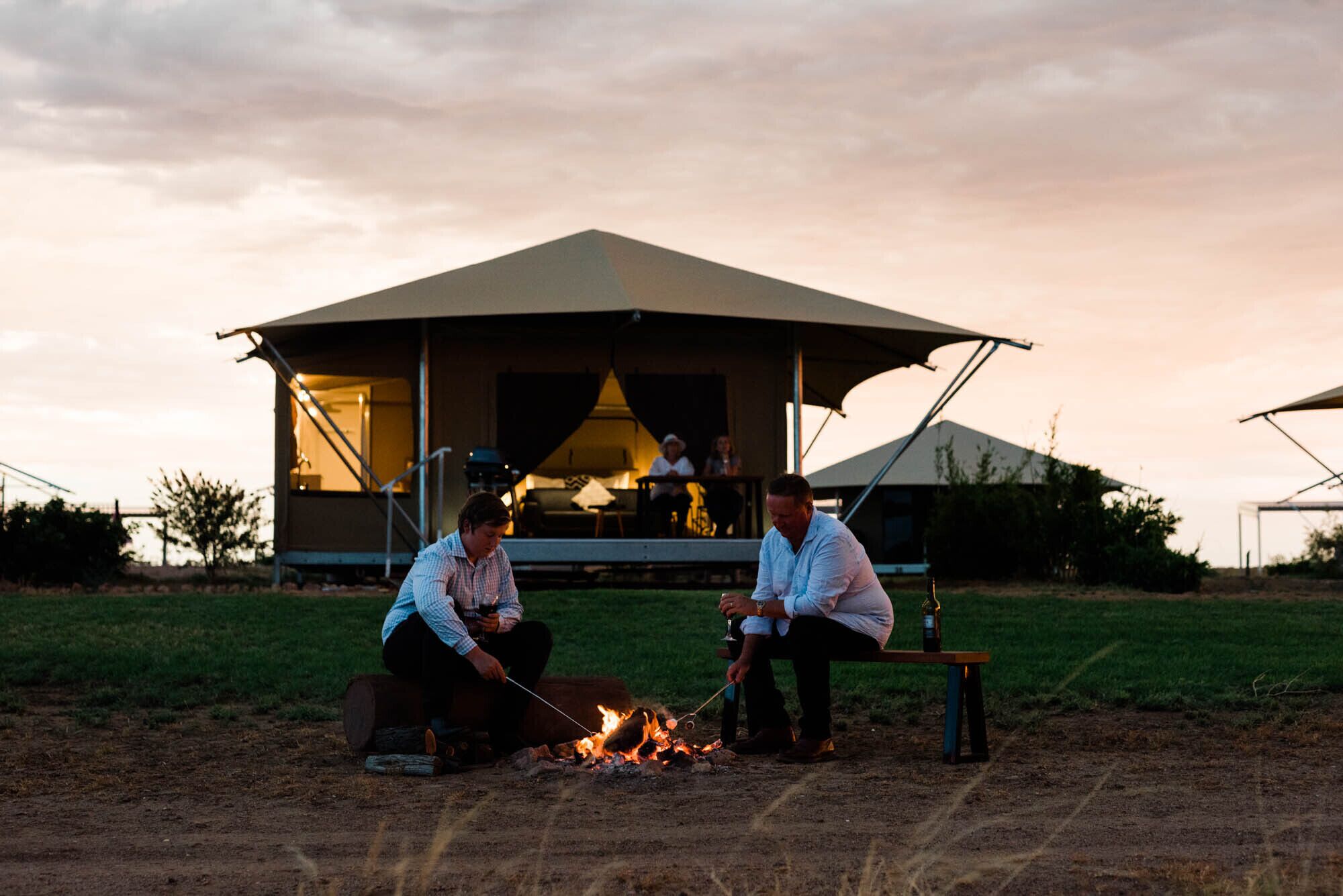 An outdoor seating area with a fire, a covered structure in the background, and a grassy field.