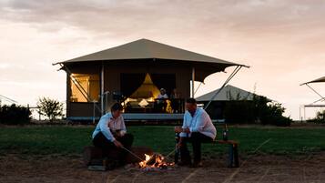 An outdoor seating area with a fire, a covered structure in the background, and a grassy field.