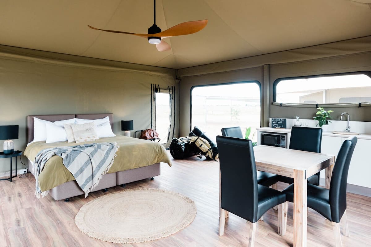A modern, well-lit room with a bed, a dining table with chairs, a kitchen area, and a ceiling fan.