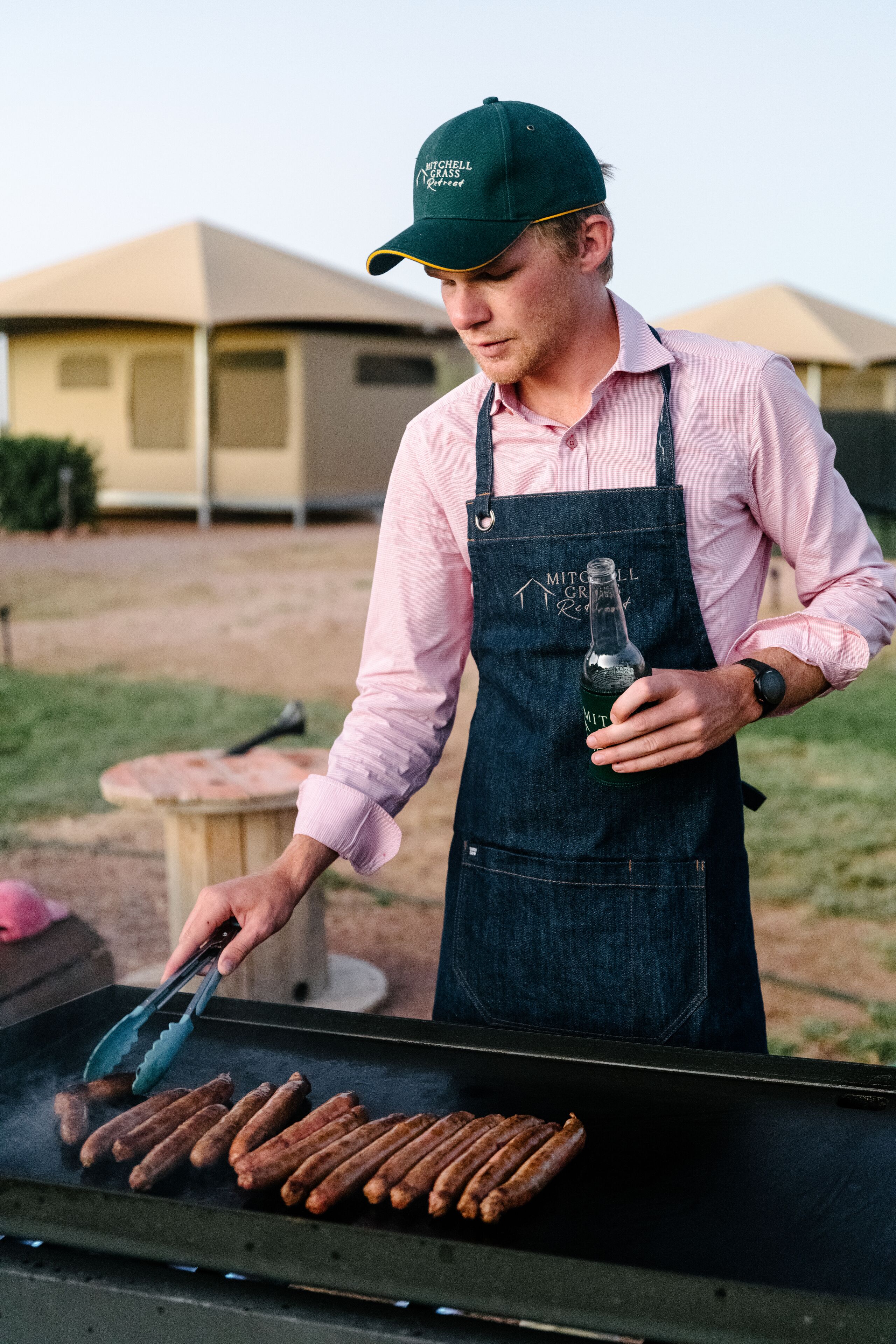 A person grilling sausages outdoors, wearing a cap and apron.