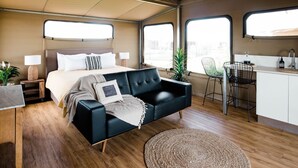 Una habitación compacta y moderna con cama, sofá negro, pequeña cocina y ventanas amplias que dejan entrar luz natural.
