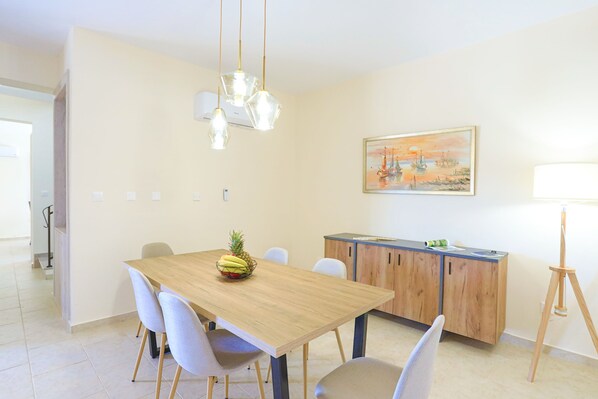 Un comedor con una mesa de madera, sillas blancas, un frutero, un aparador de madera y un cuadro en la pared.