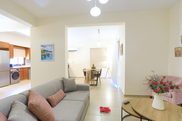 A modern living room with a grey sofa, a dining table with chairs, and a kitchen area visible in the background.