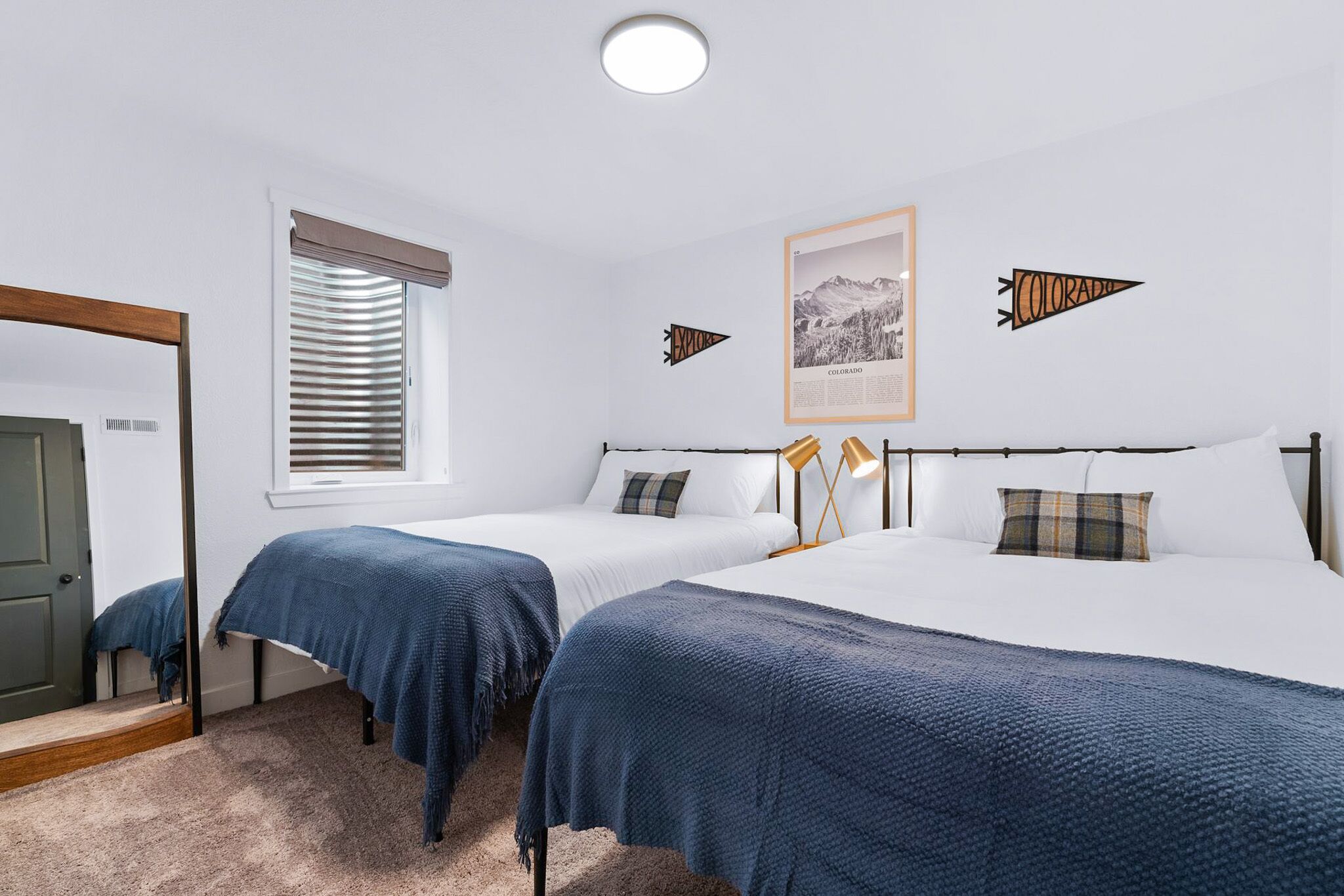 A bedroom with two beds, a window with blinds, a framed picture on the wall, and a mirror.
