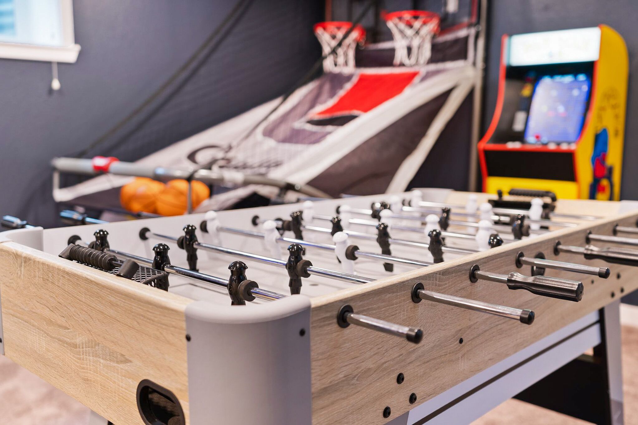 Een tafelvoetbaltafel met een houten basis en metalen stangen, in een ruimte met een basketbalring en een kleurrijke speelautomaat op de achtergrond.