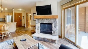 A modern living room with a stone fireplace, a flat-screen TV mounted above it, and a dining area with wooden furniture. - 1 Bedroom Mountain Condo in River Run Village With Expansive Mountain Views and Walking Distance to Ski Slopes (Keystone)