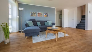 A modern living room with a sectional sofa, a coffee table, and a floor lamp. There is a staircase to the right and a door leading to another room. - Wattperle - Wunderschöne Obergeschosswohnung in Greetsiel (Krummhörn)