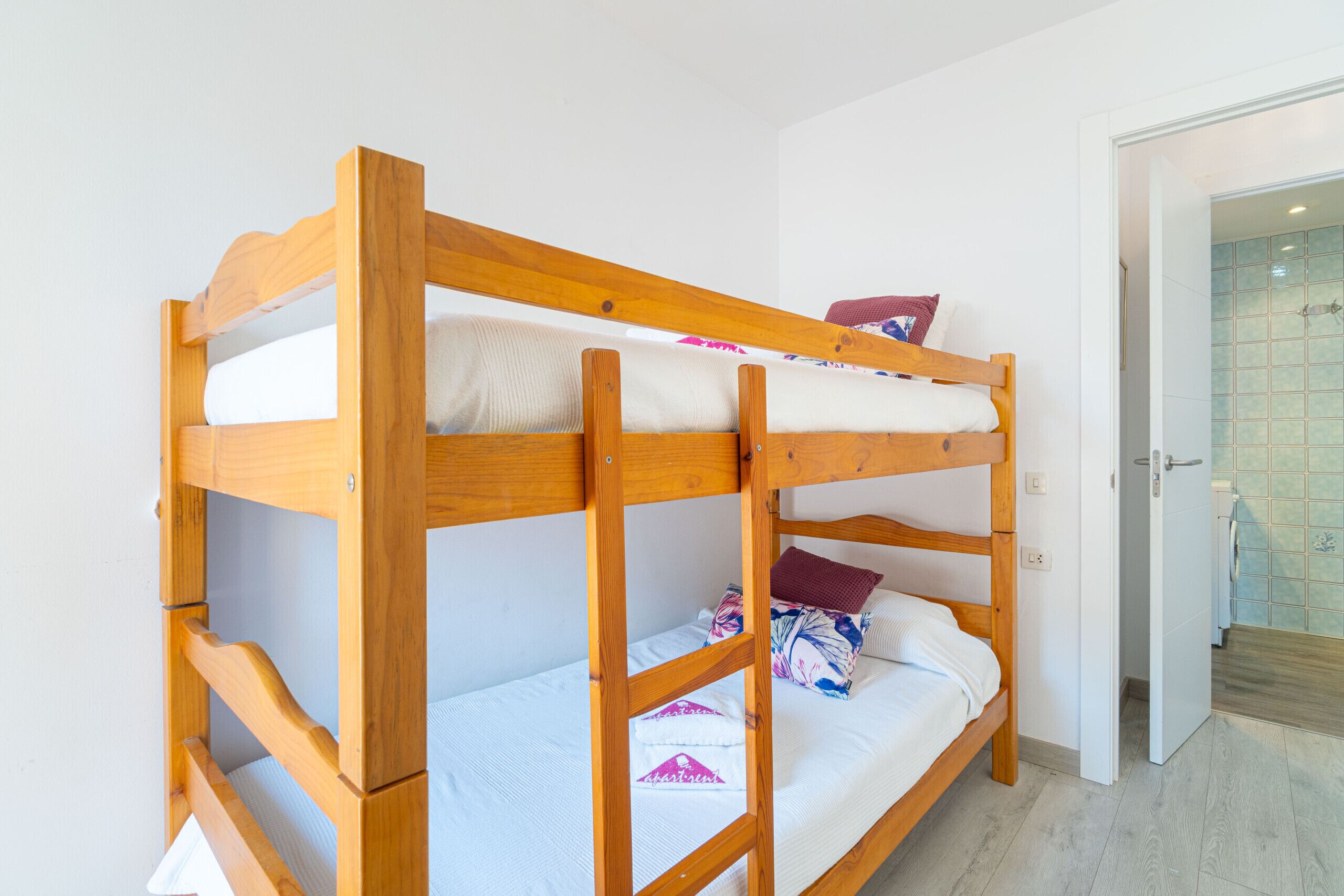 Een stapelbed met houten frame en witte beddengoed, een badkamer met betegelde douche, en een deur naar een andere kamer.