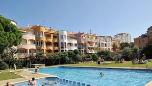 Una piscina rodeada de edificios con varios pisos y balcones, un área verde y gente nadando y descansando.