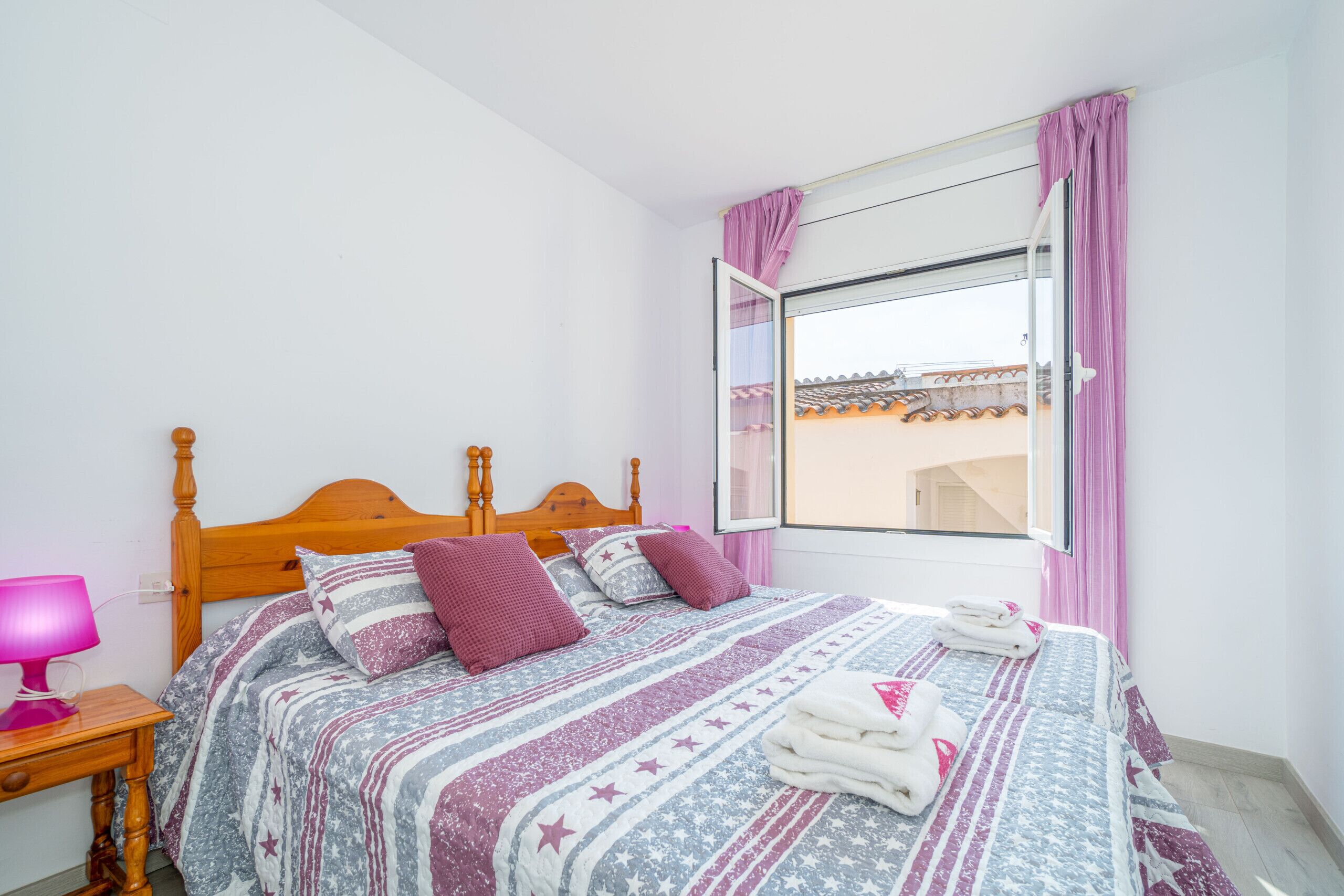 A bedroom with a wooden bed, a window with curtains, a bedside table with a lamp, and a view of buildings outside.