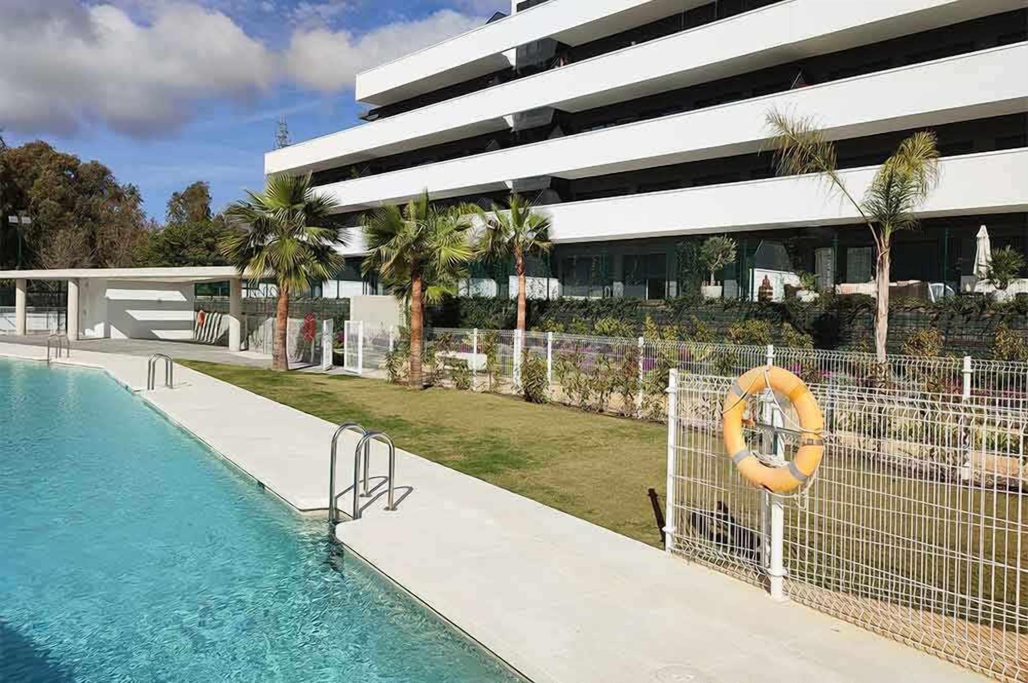 Een modern gebouw met een zwembad, palmbomen en een reddingsboei op een hek.