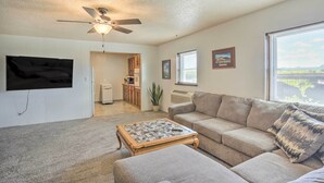 A living room with a ceiling fan, a flat-screen TV mounted on the wall, a beige sofa, a coffee table, and a window with blinds. - Peaceful Chadron Apartment in Historic Hotel! (Chadron)