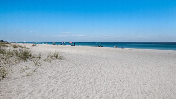 Ein weiter Sandstrand mit klarem, blauem Wasser und einigen Personen sowie Sonnenschirmen.