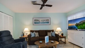 Ein Wohnzimmer mit einer Couch, zwei Beistelltischen mit Lampen, einem Couchtisch, einem Fernseher und einem Deckenventilator.