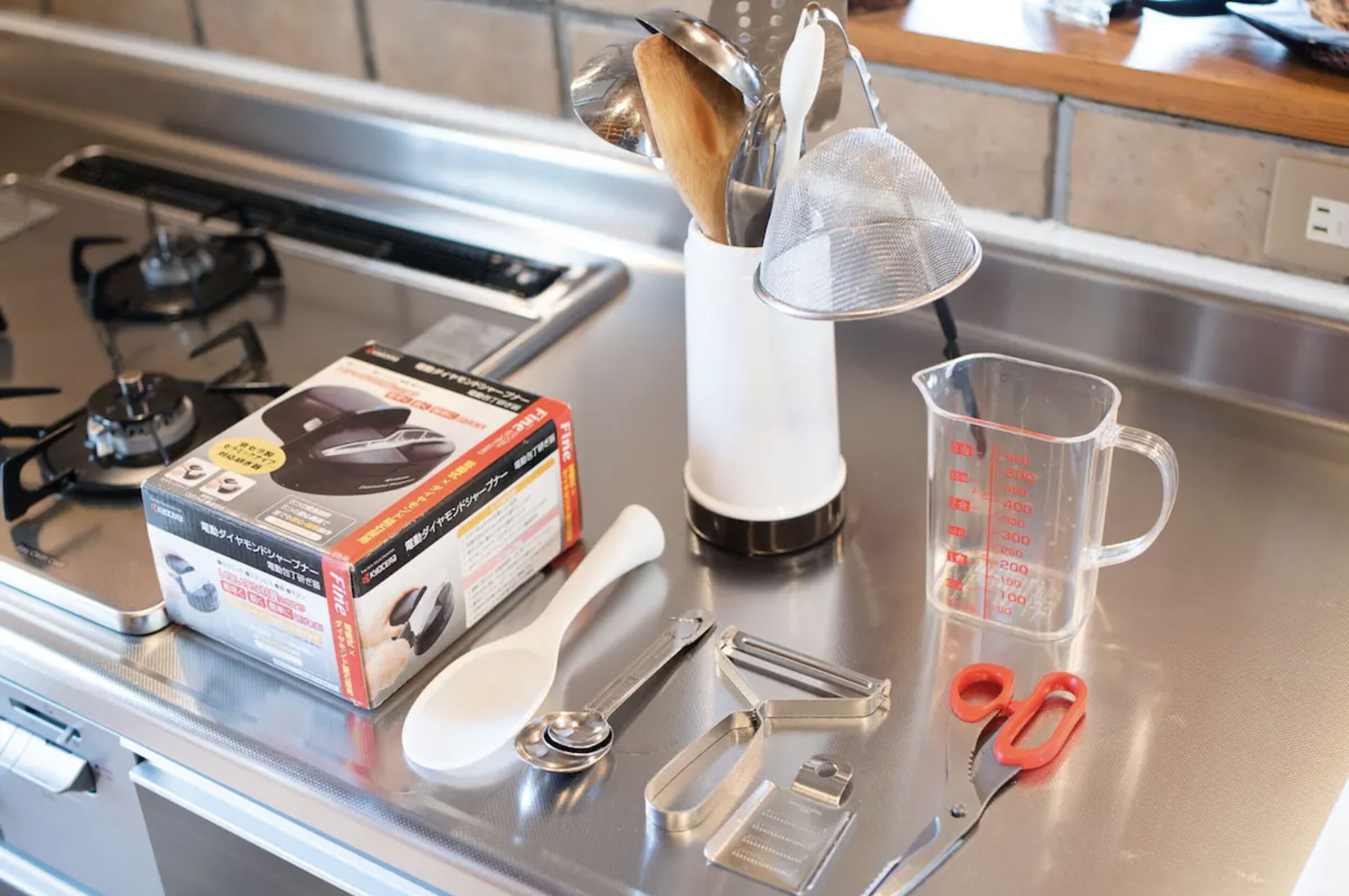A kitchen counter with a gas stove, a box of kitchen utensils, a measuring cup, and a pair of scissors.