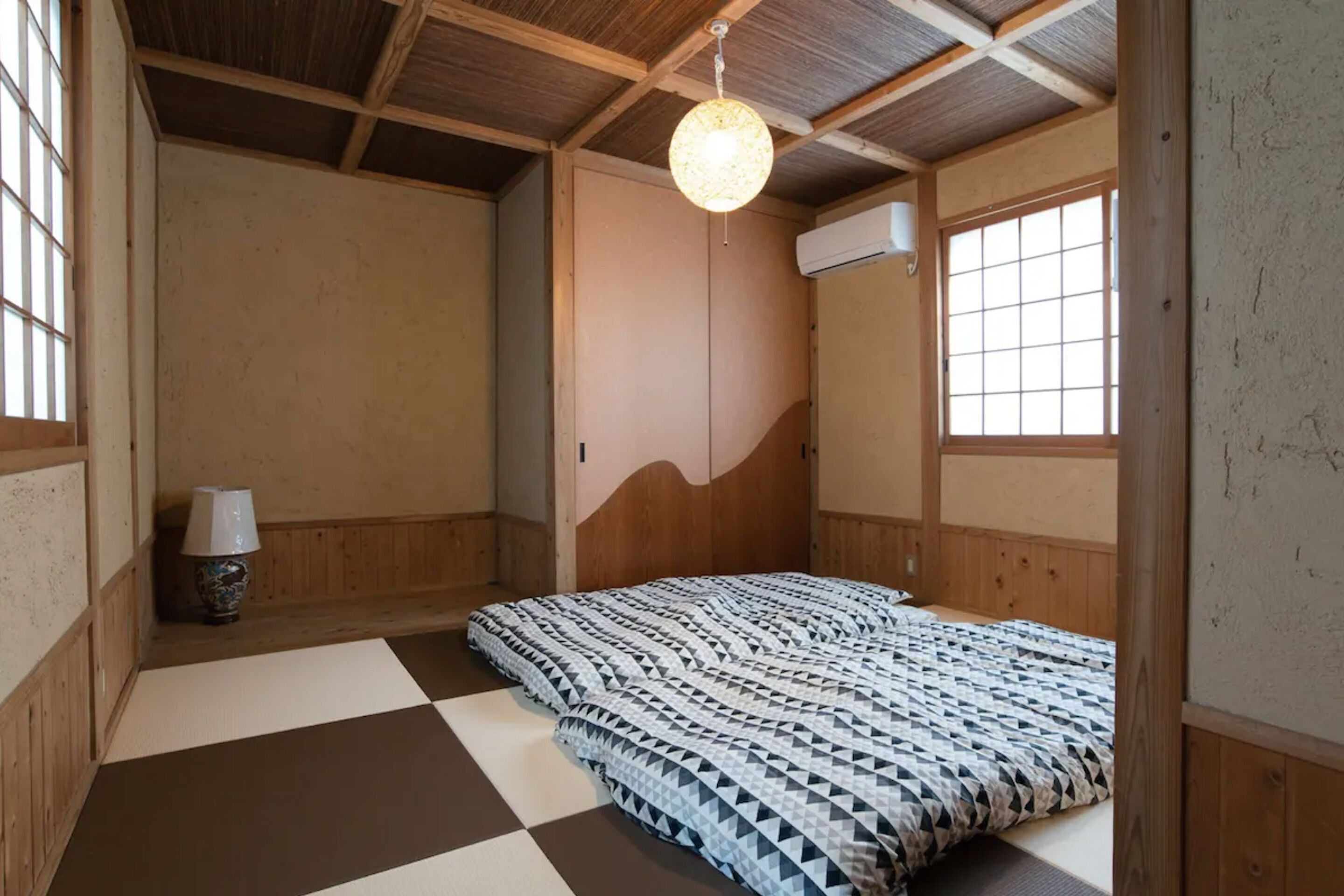 A traditional Japanese-style room with tatami flooring, a bed with a patterned bedspread, a hanging light fixture, and a sliding door.