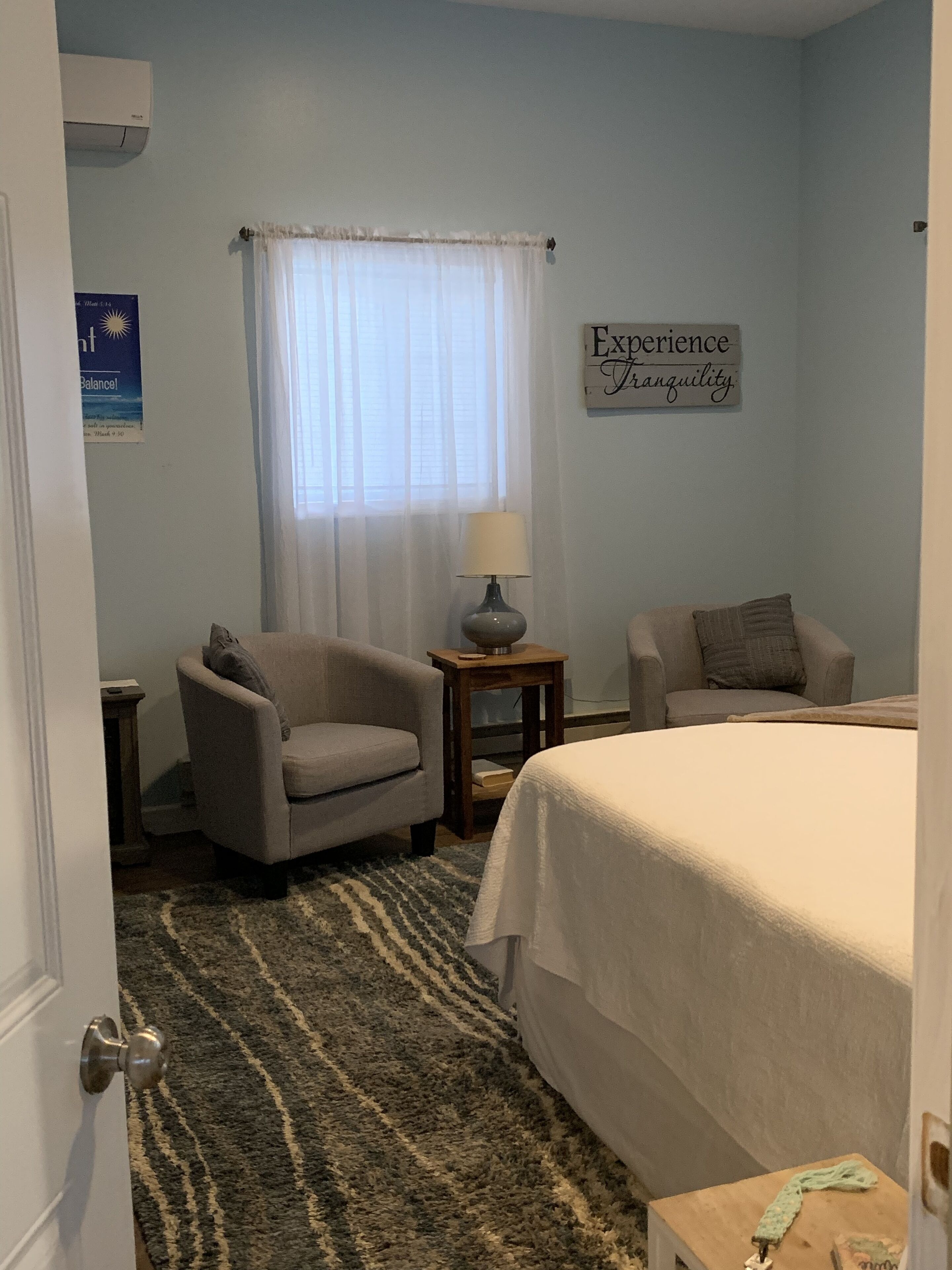 A bedroom with a bed, two armchairs, a small table with a lamp, a window with sheer curtains, and a wall sign.