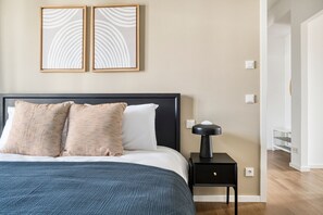 Quarto com uma cama, duas almofadas, uma mesinha de cabeceira com abajur e duas obras de arte na parede.