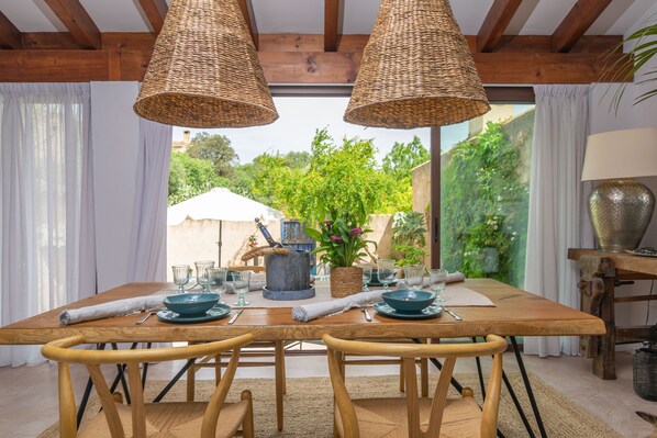 A dining area with a wooden table set for two, surrounded by wicker lamps, and a view of greenery outside.