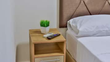 Een net opgemaakt bed met witte lakens, een nachtkastje met een afstandsbediening, een boek en een kleine potplant.