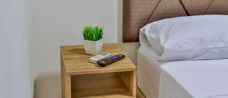 Una cama bien hecha con sábanas blancas, una mesita de noche con un control remoto, un libro y una pequeña planta en maceta.