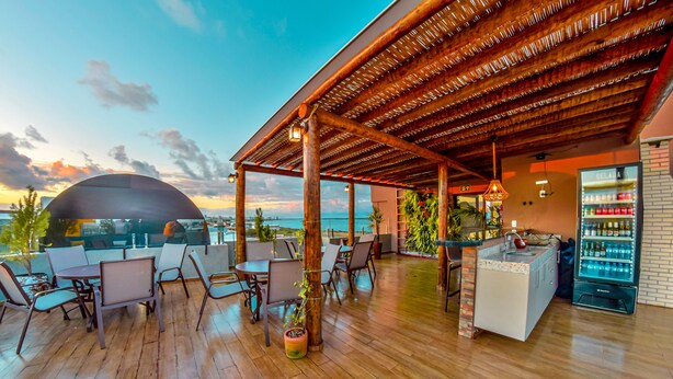 Un espacio al aire libre con piso de madera, mesas, sillas y una zona de bar con refrigerador.